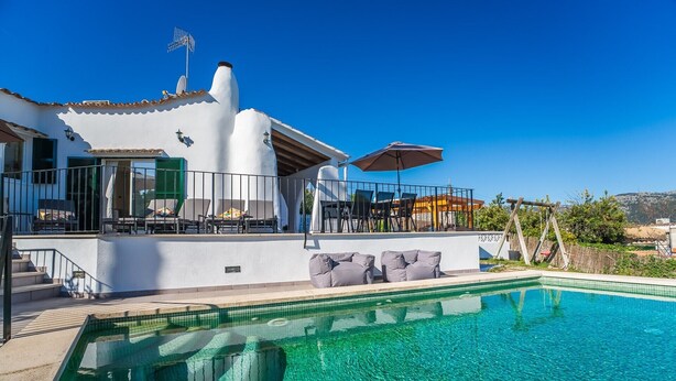 Un espace aménagé au bord d’une piscine à l’eau d’un bleu limpide, avec des sièges d’extérieur et un bâtiment blanc doté d’une cheminée remarquable.