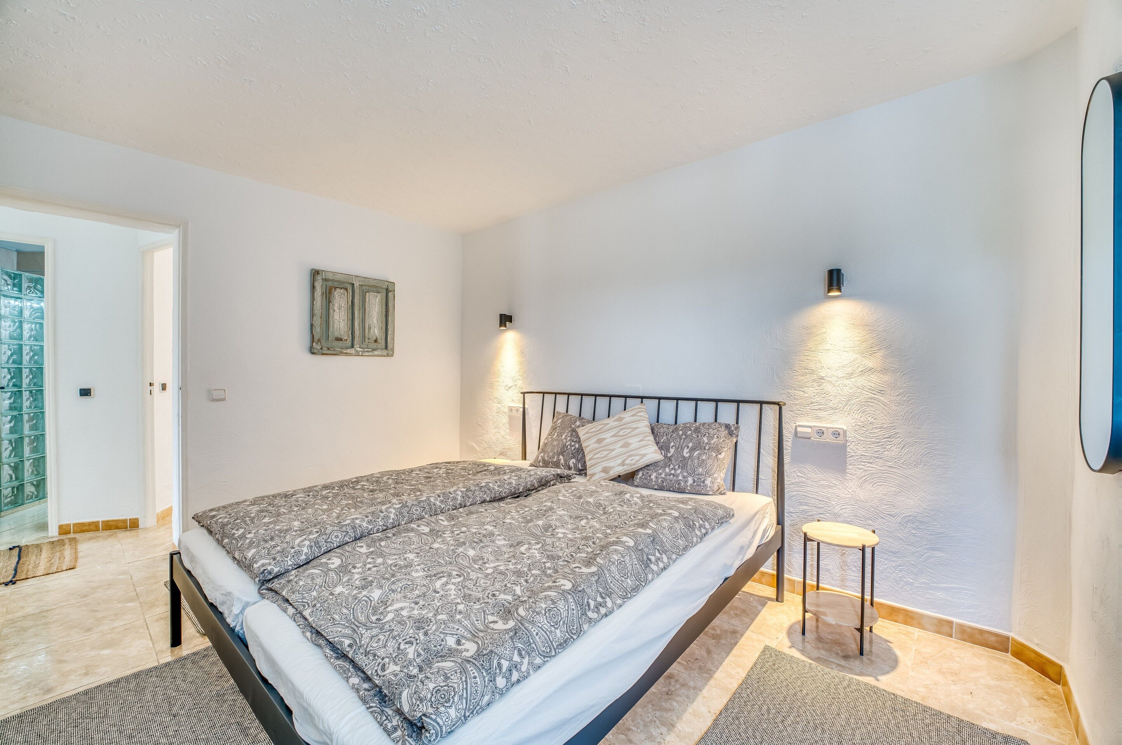 A bedroom with a bed, two wall-mounted lights, a small stool, and a door leading to a balcony.