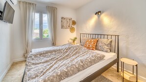Une chambre à coucher avec un lit, des rideaux, un téléviseur fixé au mur, une table de chevet avec une plante et un luminaire fixé au mur.