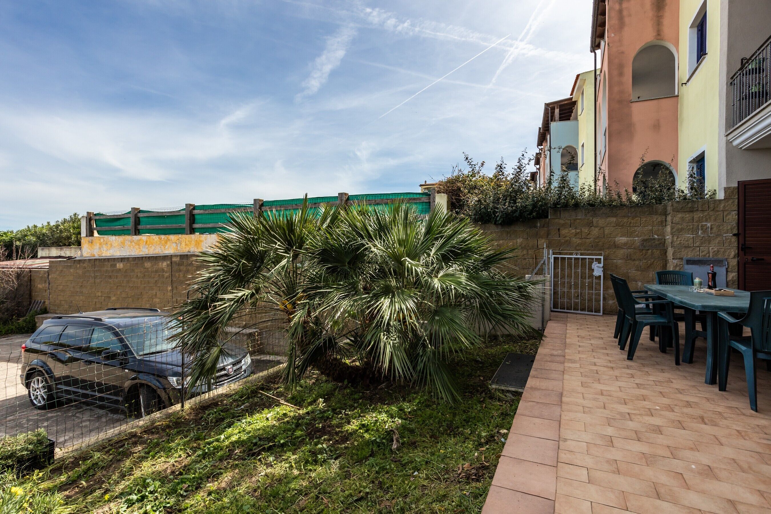 A courtyard with a car parked by a fence, a palm tree, and a table with chairs.
