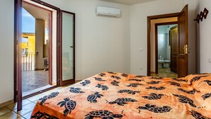 A bedroom with a bed, an air conditioning unit, a sliding glass door, and a bathroom visible through an open door.
