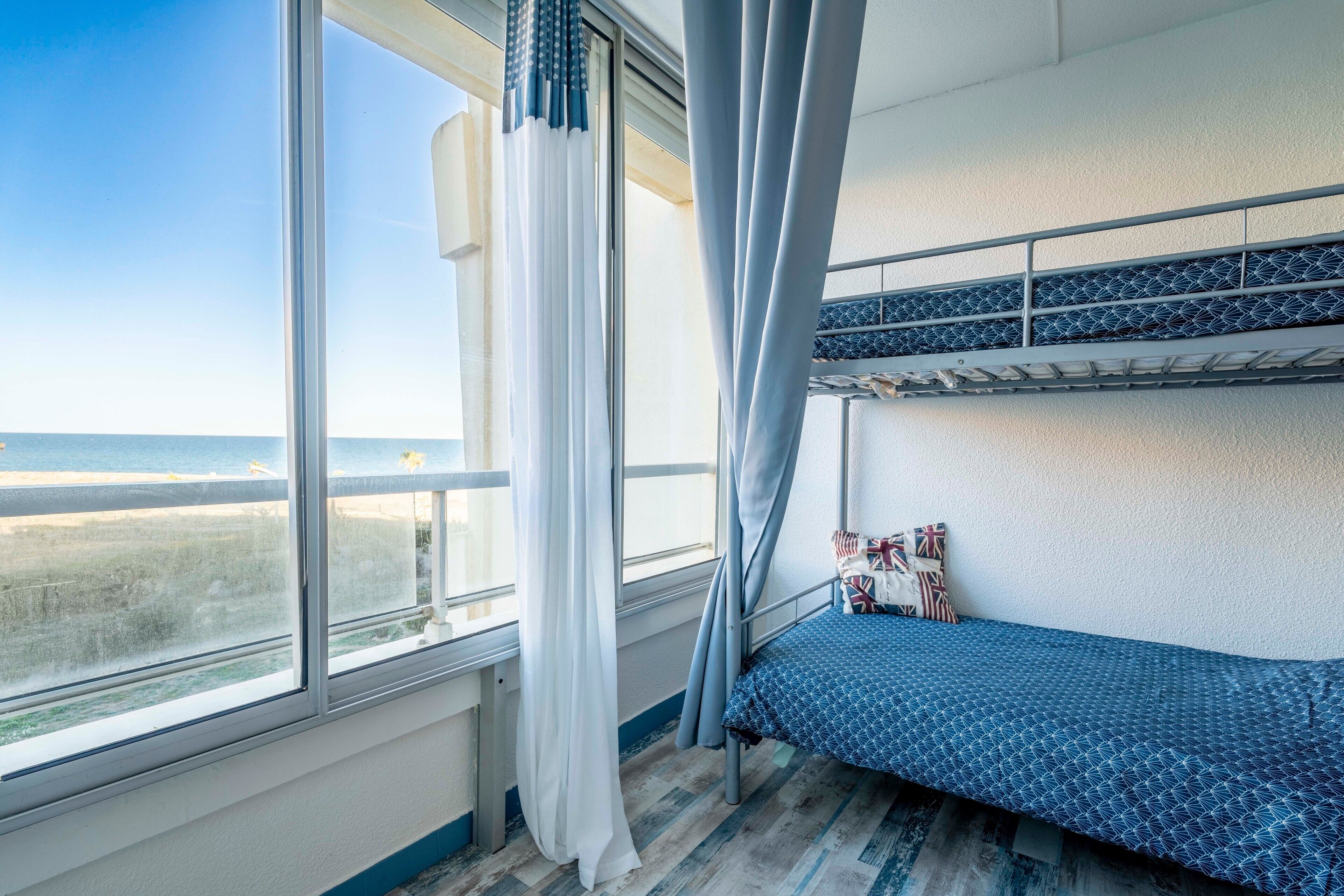 Ein Schlafraum mit Etagenbetten und Blick auf den Strand, ein Fenster mit Vorhängen und einer gemusterten Bettdecke.