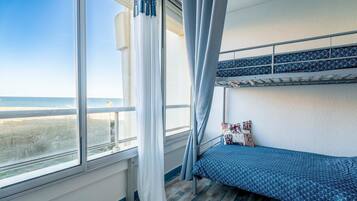 Ein Schlafraum mit Etagenbetten und Blick auf den Strand, ein Fenster mit Vorhängen und einer gemusterten Bettdecke.