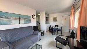 A living room with a sofa, a coffee table, a dining area with chairs, a kitchen in the background, and a door. - Apartment 'Blaumar B310' with Shared Pool, Private Terrace and Wi-Fi (LÈstartit)