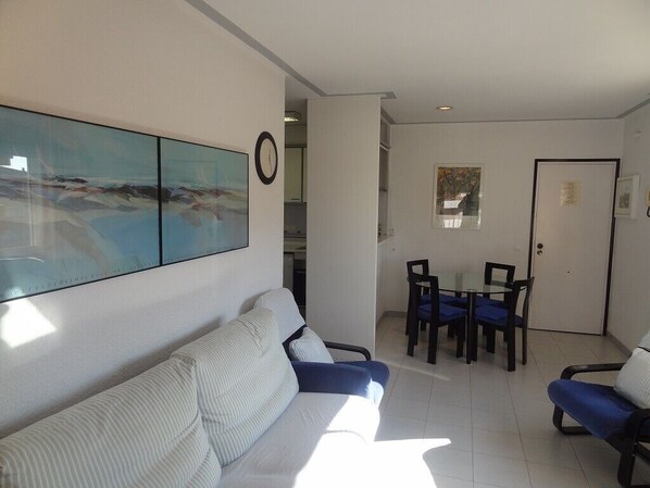A living room with a sofa, a dining table with chairs, a clock on the wall, and a door.