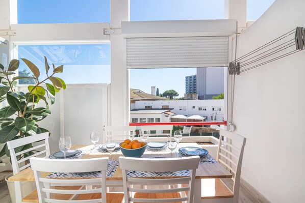 A dining area with a table set for two, a bowl of fruit, and a view of buildings through a window.