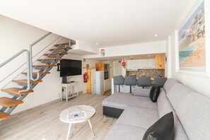 A modern living room with a grey sofa, wooden staircase, flat-screen TV, and a small dining area with chairs.