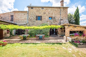 Un edificio de piedra con un patio jardín, una mesa con sillas y una pequeña construcción adornada con flores.