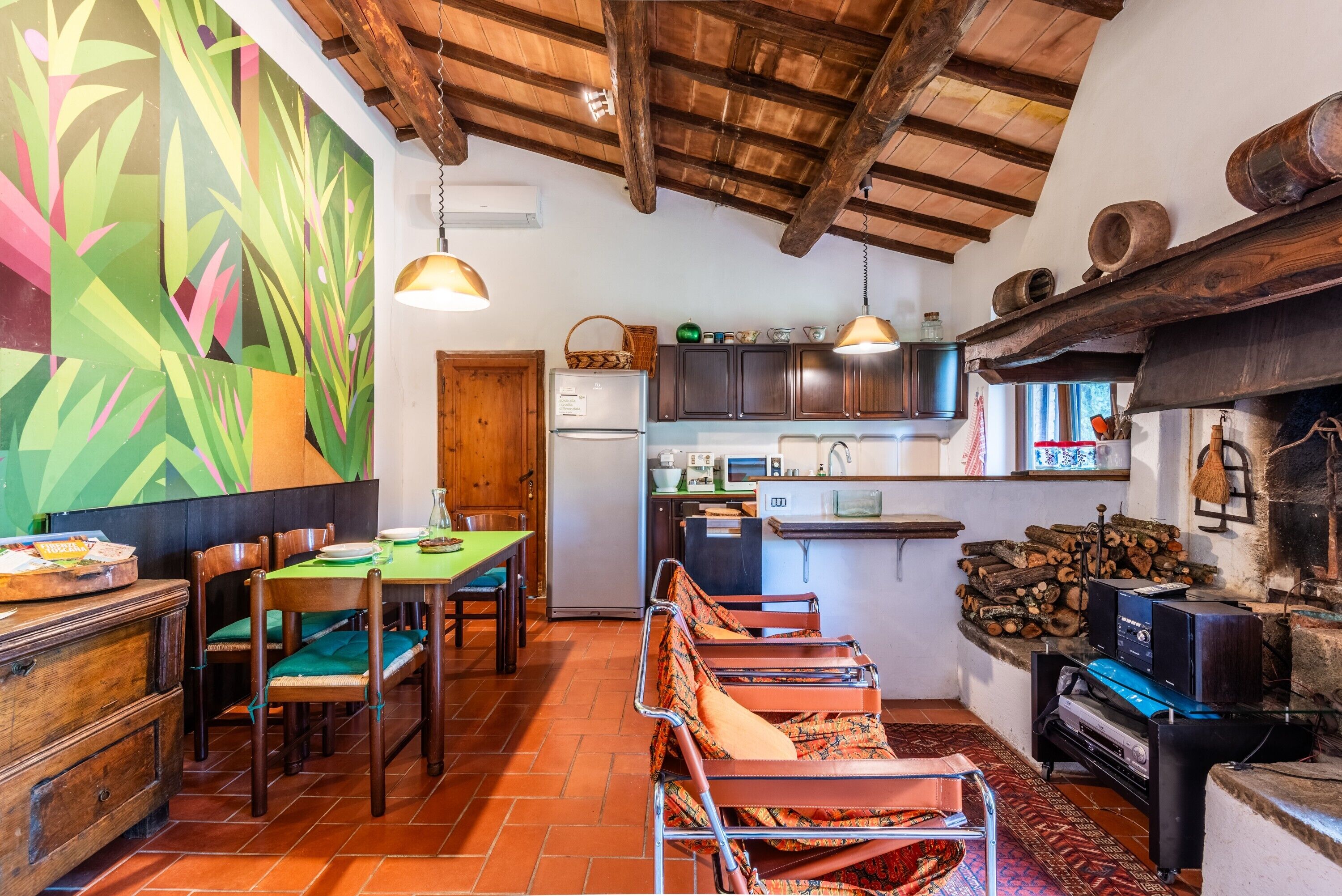 A rustic kitchen with wooden beams, a dining area with chairs, a colorful wall, and a fireplace.