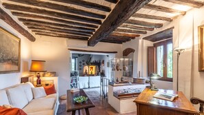 Ein gemütliches Wohnzimmer mit einer Couch, einem Holztisch und einem Schreibtisch mit Lampe. Im Hintergrund ist eine Küchenzeile mit Esstisch und Stühlen zu sehen.