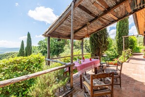 Un área de estar al aire libre techada, con una mesa y sillas, rodeada de abundante vegetación y vistas al campo.