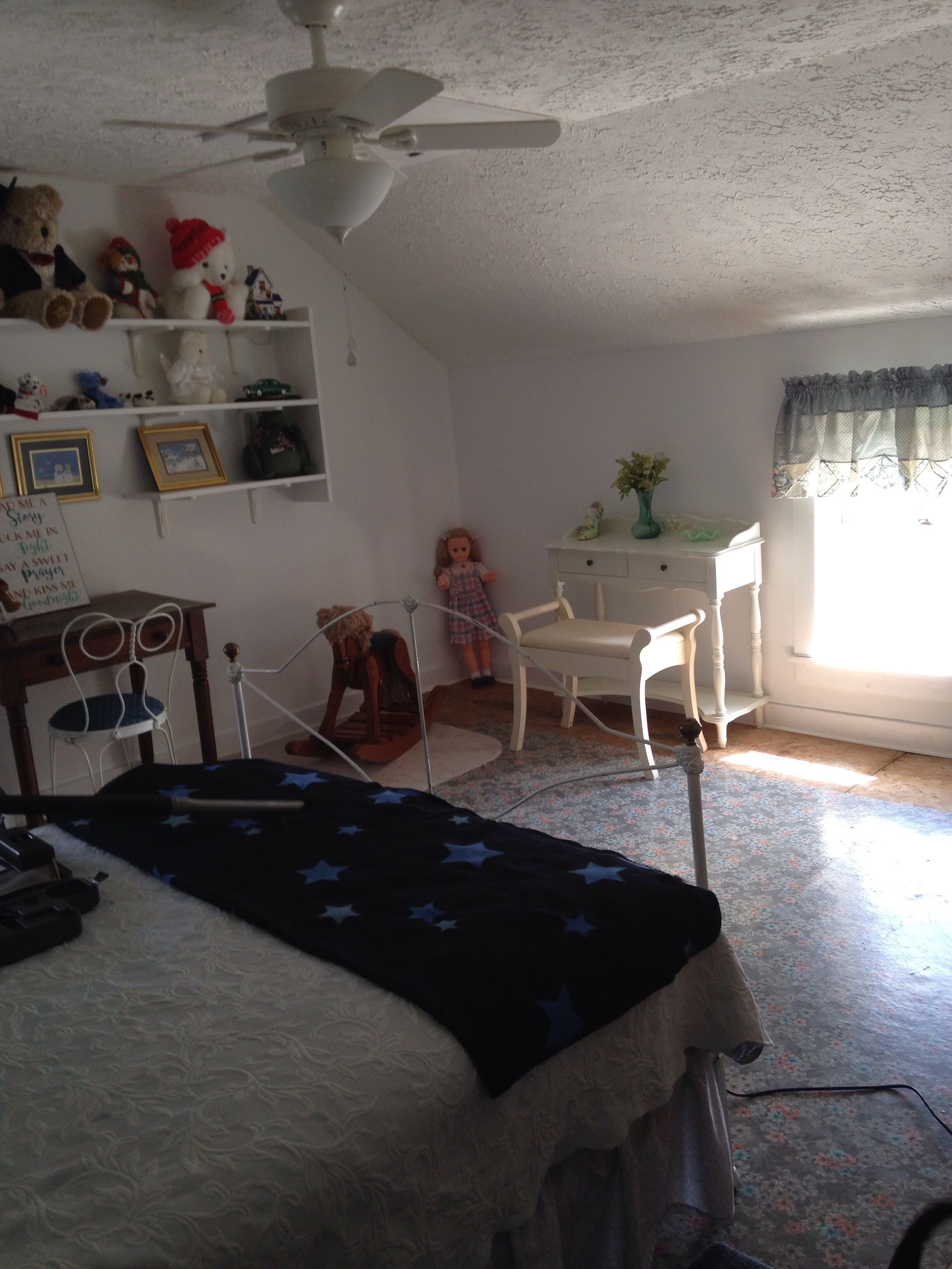 A bedroom with a bed, a desk, a chair, a rocking chair, a ceiling fan, and a window with curtains.