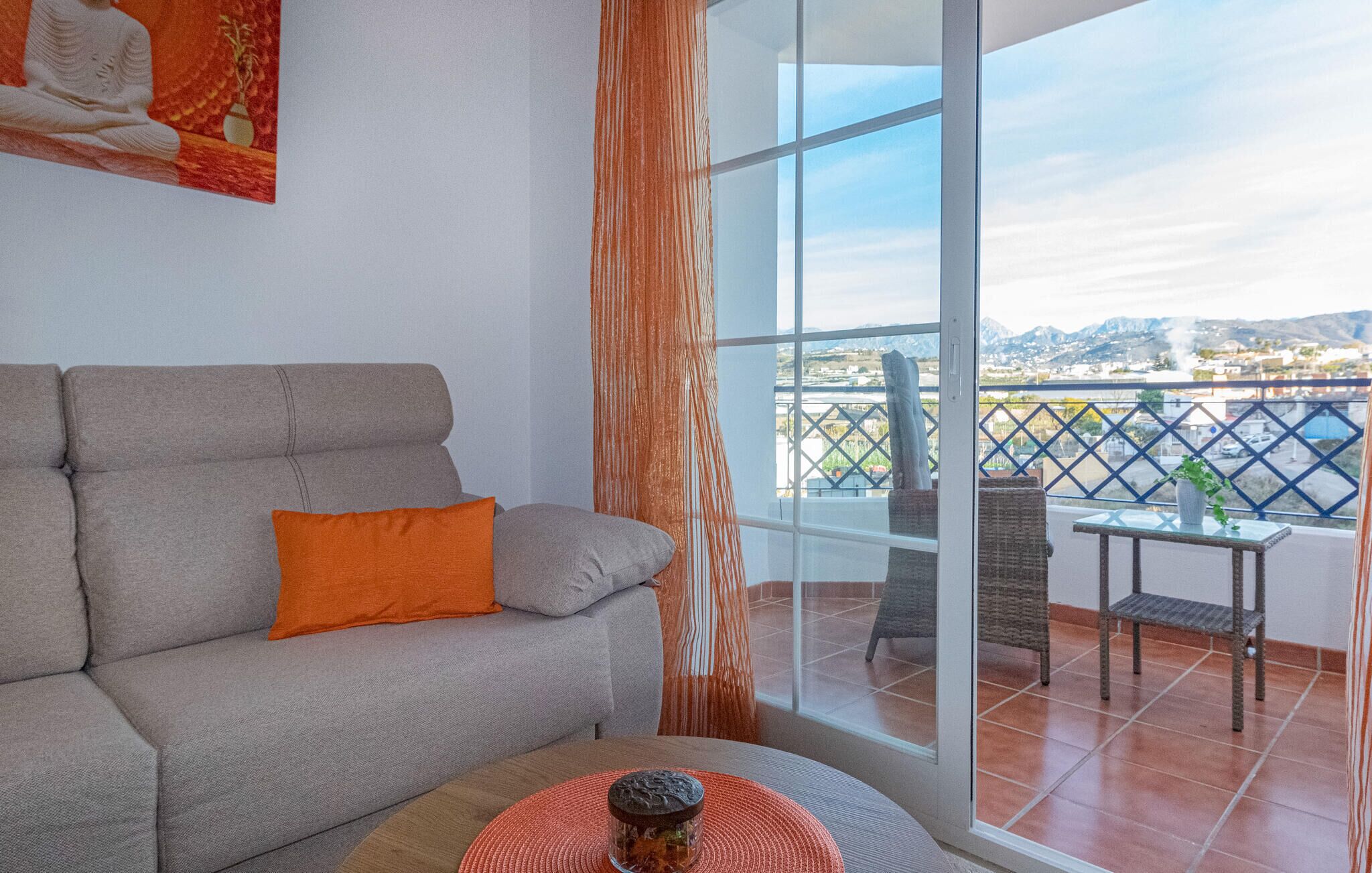 A living room with a grey sofa, an orange cushion, a glass table, and a view of a city and mountains through large windows.