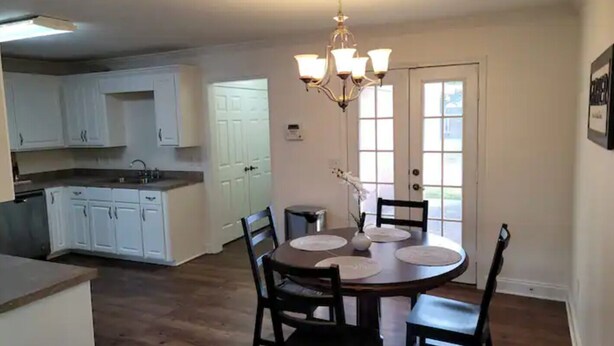 A dining area with a round table, chairs, and a chandelier. There is a kitchen with white cabinets and a window.