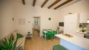 Una cuina moderna amb armaris blancs, una zona de menjador amb cadires verdes i un saló amb sofà i una planta.