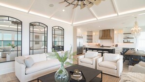 A spacious living area with a large sofa, a coffee table, and a dining table. There is a kitchen in the background with modern appliances and a chandelier. - Hobbs Realty - Luxury Oceanfront Home w/Heated Pool, Elevator, and Media Room (Holden Beach)