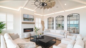 A spacious living room with a modern fireplace, white sofas, a black coffee table, and large windows offering a view of the sea. - Hobbs Realty - Luxury Oceanfront Home w/Heated Pool, Elevator, and Media Room (Holden Beach)