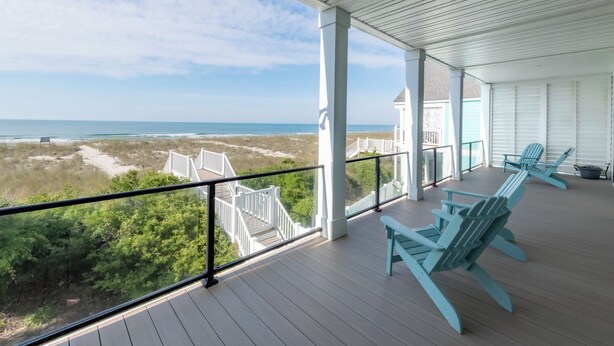 A covered balcony with wooden flooring, white railings, and turquoise Adirondack chairs. - Hobbs Realty - Luxury Oceanfront Home w/Heated Pool, Elevator, and Media Room (Holden Beach)