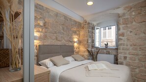 A bedroom with a stone wall, a bed with a beige headboard, a bedside table with a lamp, and a window with shutters. - MIRABILIS - studio apartment - Old Town (Dubrovnik)