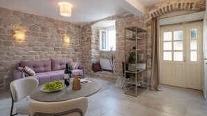 A cozy living area with a stone wall, a purple sofa, a round table with a bottle and a bowl of fruit, and a bookshelf. - MIRABILIS - studio apartment - Old Town (Dubrovnik)