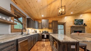 A modern kitchen with wooden cabinets, a large island, and a stone fireplace. - JULY DISCOUNT! New Modern Luxury Cabin- Hot Tub, Game Room, Fire Pit (Blue Ridge)