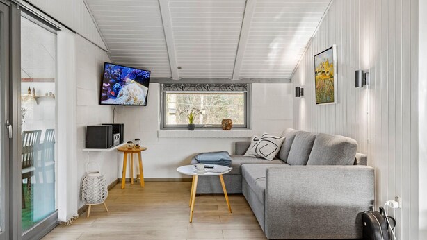 Ein modernes Wohnzimmer mit einem grauen Sofa, einem an der Wand montierten Fernseher und einem Fenster mit Blick auf Bäume.