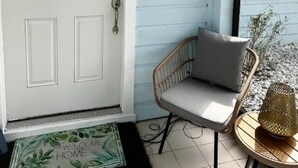 A white door with a doormat, a wicker chair with a cushion, a small table with a lamp, and a wall-mounted light fixture. - A Peaceful Family Friendly Home in Sarasota Fla, Legacy Trail/Pinecraft (Sarasota)