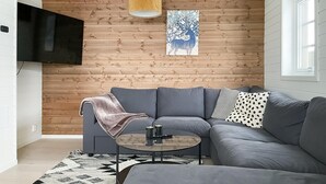 Una sala de estar moderna con un sofá seccional gris, una mesa de centro redonda con controles remotos, un plaid y una pared de madera con un televisor montado y una obra de arte enmarcada.