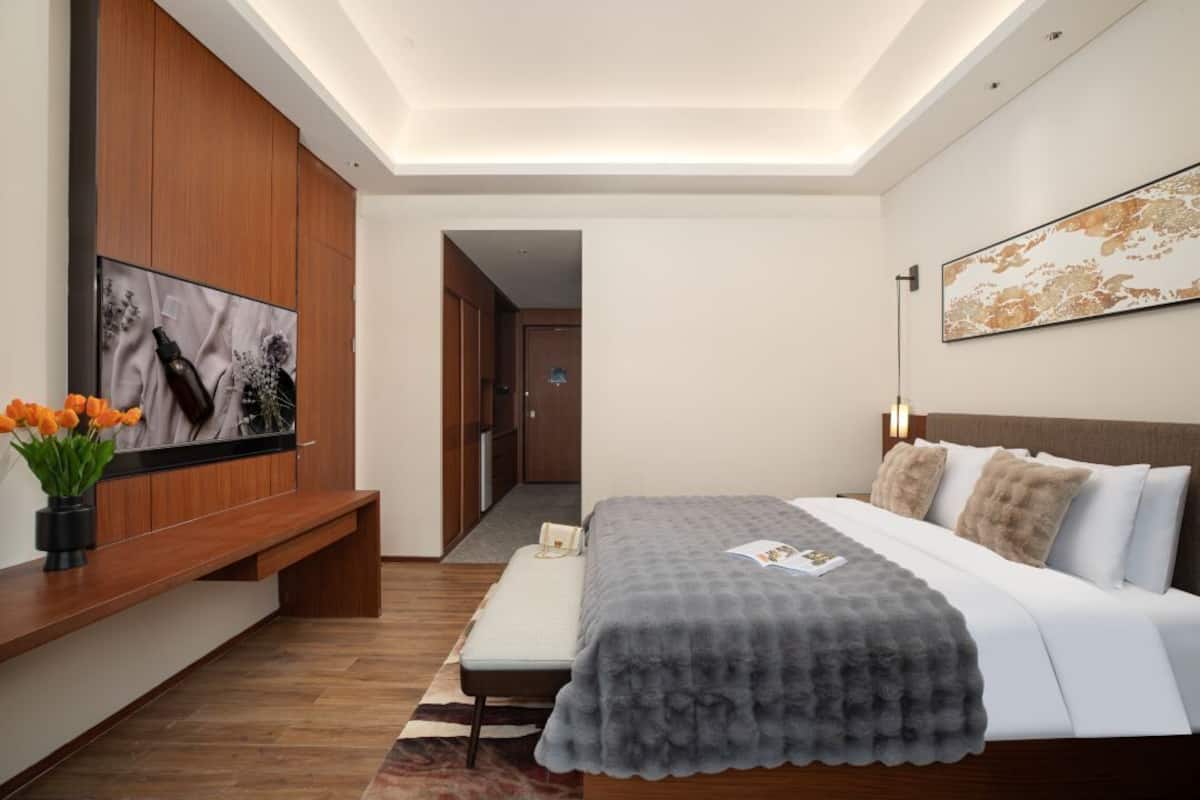 A modern hotel room with a large bed, a flat-screen TV mounted on a wooden panel, a bench with a vase of flowers, and a hallway leading to another room.