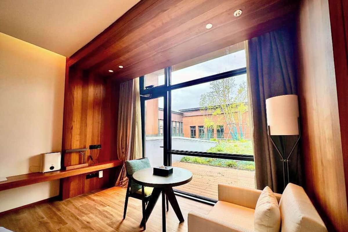 A hotel room with a wooden ceiling, a large window with a view of the outside, a round table, and a sofa.
