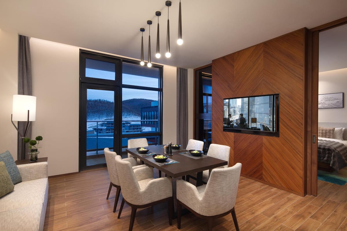 A modern hotel room with a dining area, large windows with a view of a snowy landscape, a wooden accent wall, and a flat-screen TV mounted on the wall.