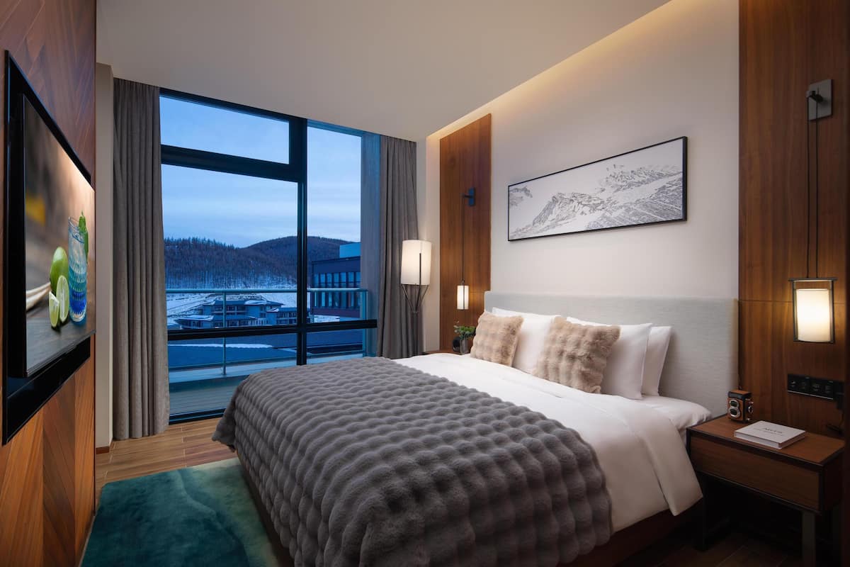 A modern hotel room with a large bed, a view of a snowy landscape, a mounted painting, and a bedside table with a lamp.