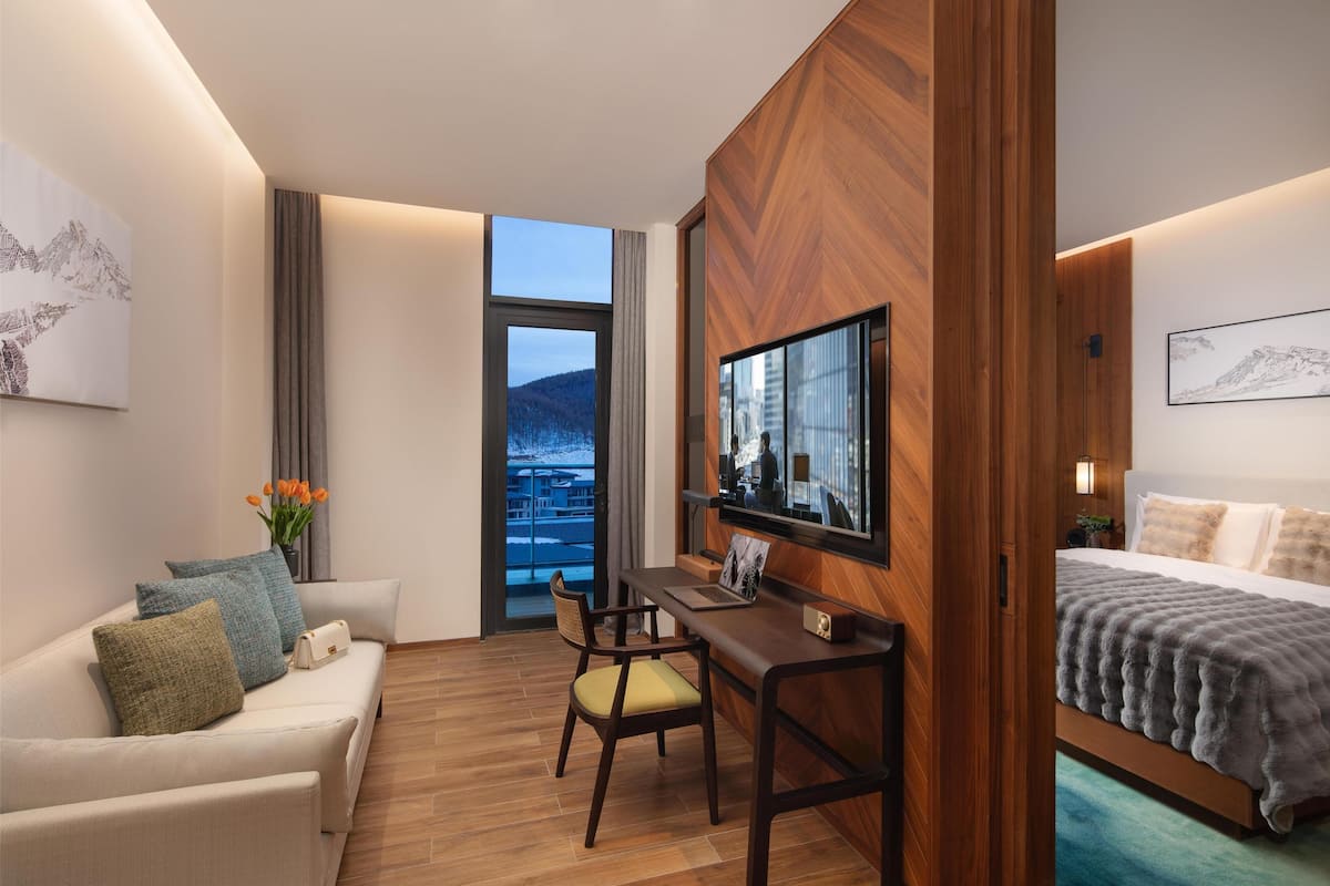 A modern hotel room with a wooden desk, a comfortable sofa, a bed, and a large window offering a view of a mountain and cityscape.