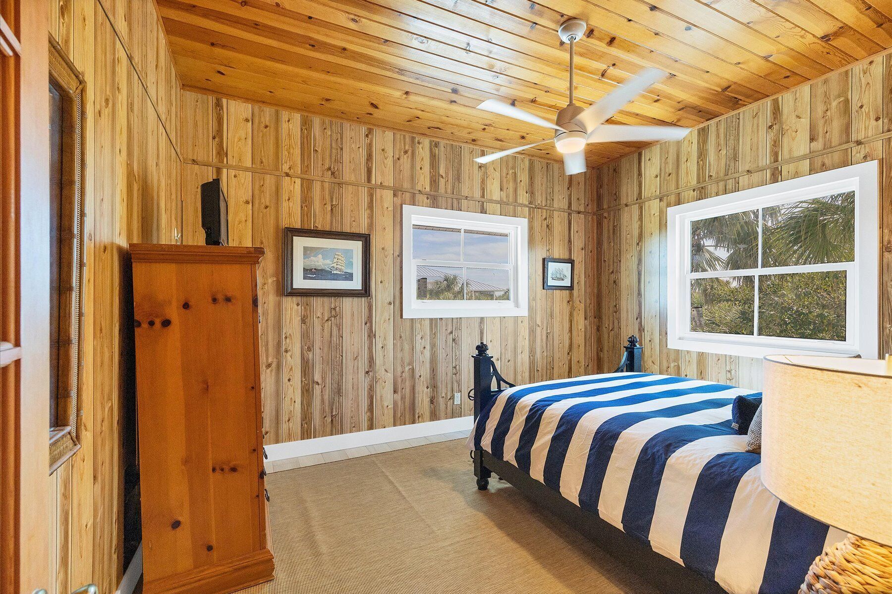 A bedroom with wooden walls, a bed with striped bedding, a ceiling fan, and two windows.