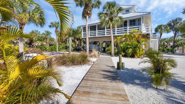 Ein modernes Strandhaus mit einer Holzterrasse, die zum Eingang führt, umgeben von Palmen und einem klaren Himmel.