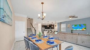 A dining area with a wooden table set for six, a chandelier, and a view of the kitchen and living area.
