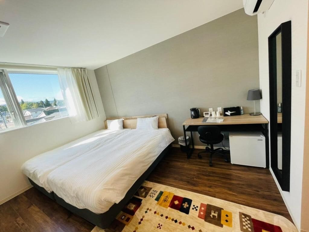 Un dormitorio con cama, escritorio, silla, un refrigerador pequeño y ventana con vistas a edificios.