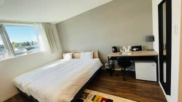 Un dormitorio con cama, escritorio, silla, un refrigerador pequeño y ventana con vistas a edificios.