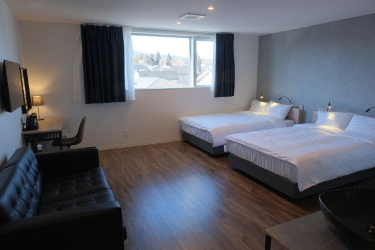 A hotel room with two single beds, a sofa, a desk with a lamp, and a view of the city.