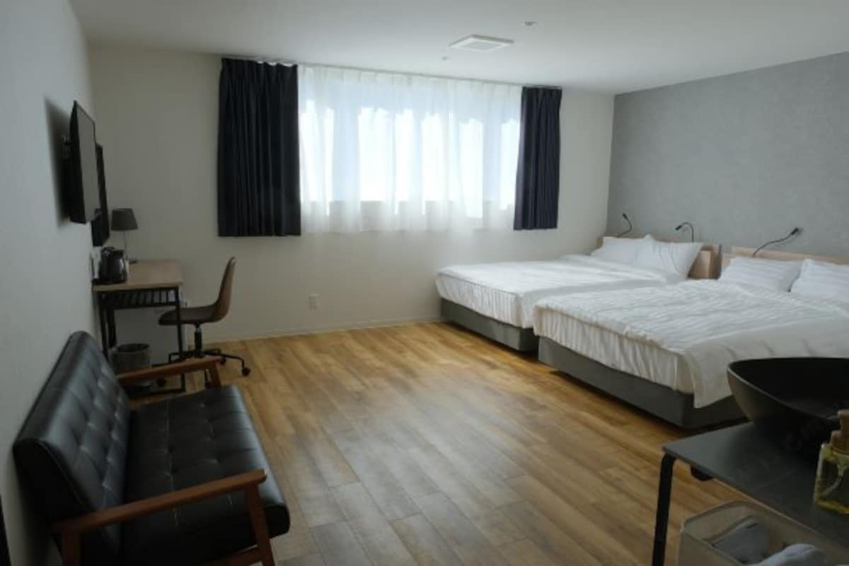A modern hotel room with a wooden floor, a sofa, a desk with a chair, two beds, and a large window with curtains.