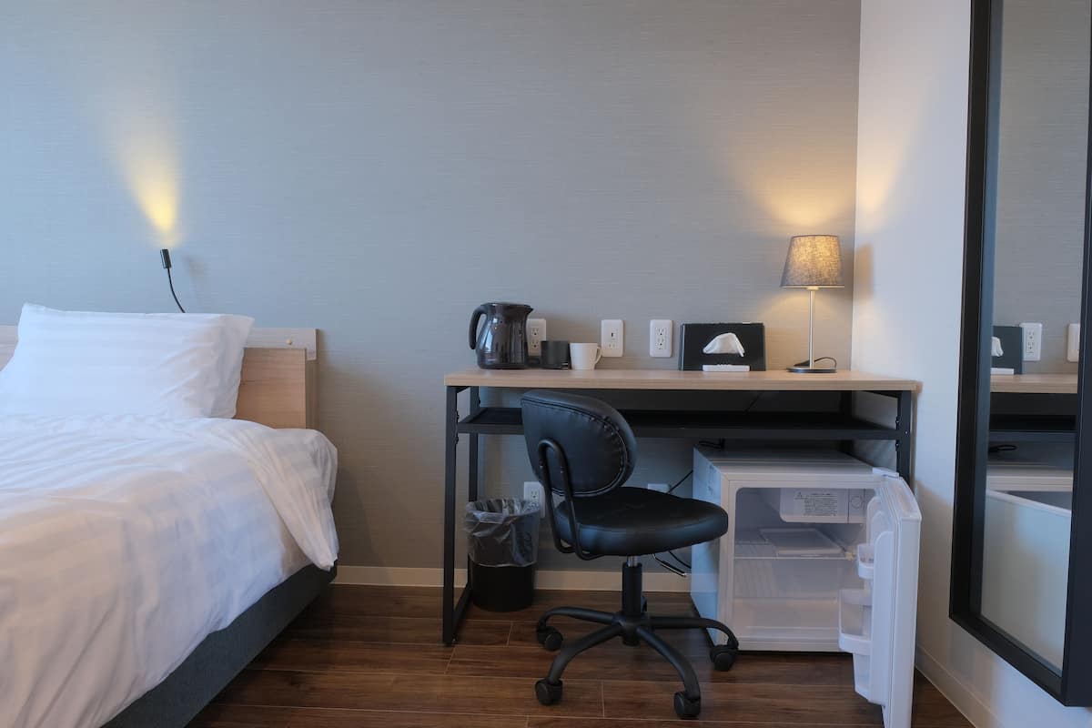 Una habitación de hotel con cama, escritorio con silla, lámpara, cafetera y basurero.