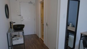 Une petite pièce aux dimensions réduites, avec un plancher en bois, une porte blanche, un bol noir posé sur une étagère en métal, un miroir au mur et un petit réfrigérateur blanc.