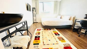 A hotel room with a bed, a desk with a chair, a TV, a window with curtains, and a patterned rug.