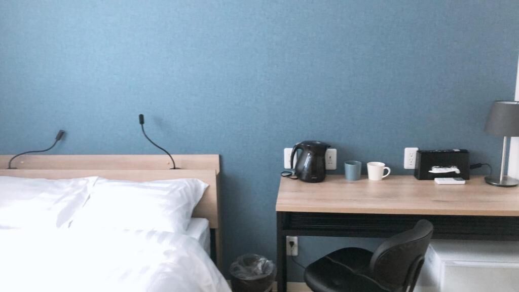 A hotel room with a bed, a desk with a coffee maker, a trash bin, and a chair.