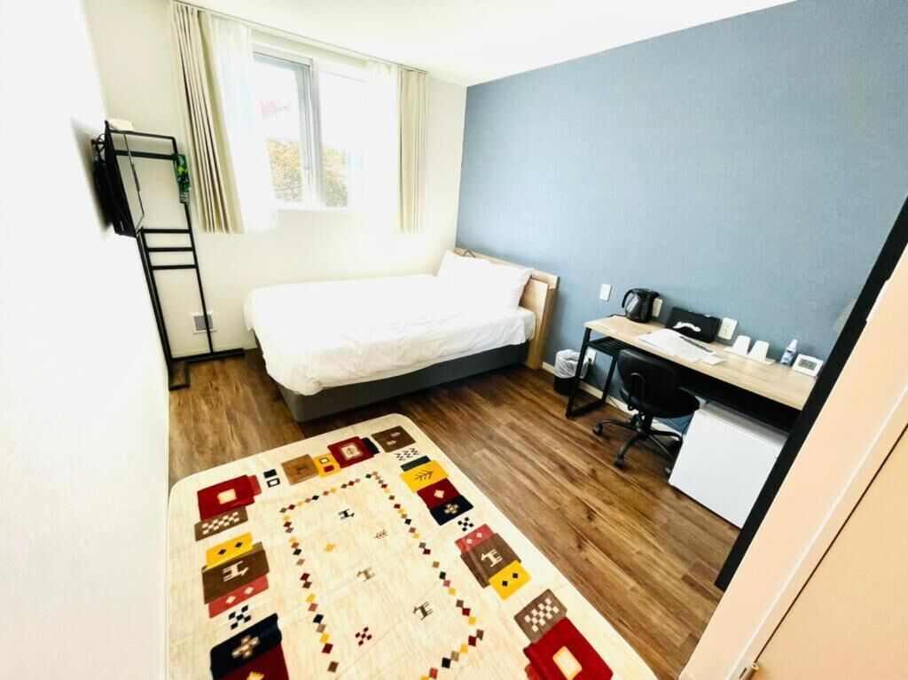 A bedroom with a bed, desk, and chair, a small kitchenette, and a window with curtains.