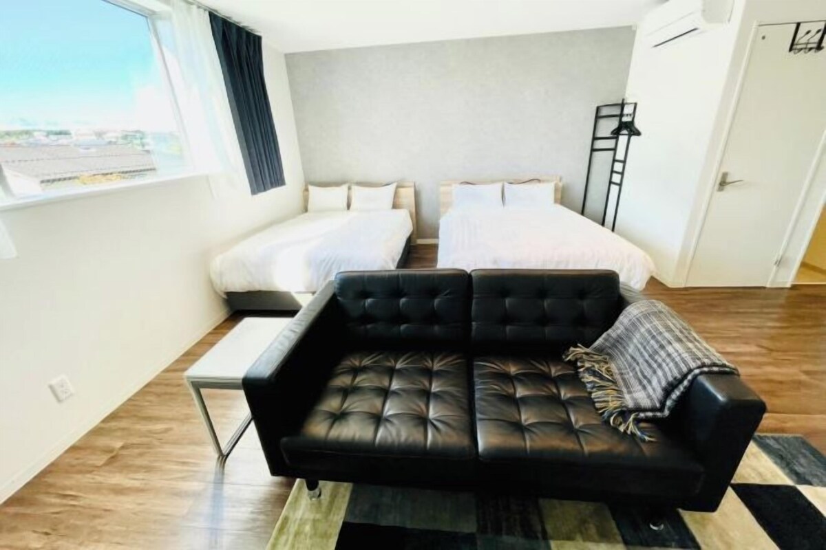 A modern hotel room with a black leather sofa, two single beds, a small table, and a view of the city through the window.