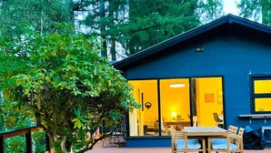 Une terrasse en bois avec une table et des chaises, un grand arbre et un bâtiment bleu doté de grandes fenêtres.