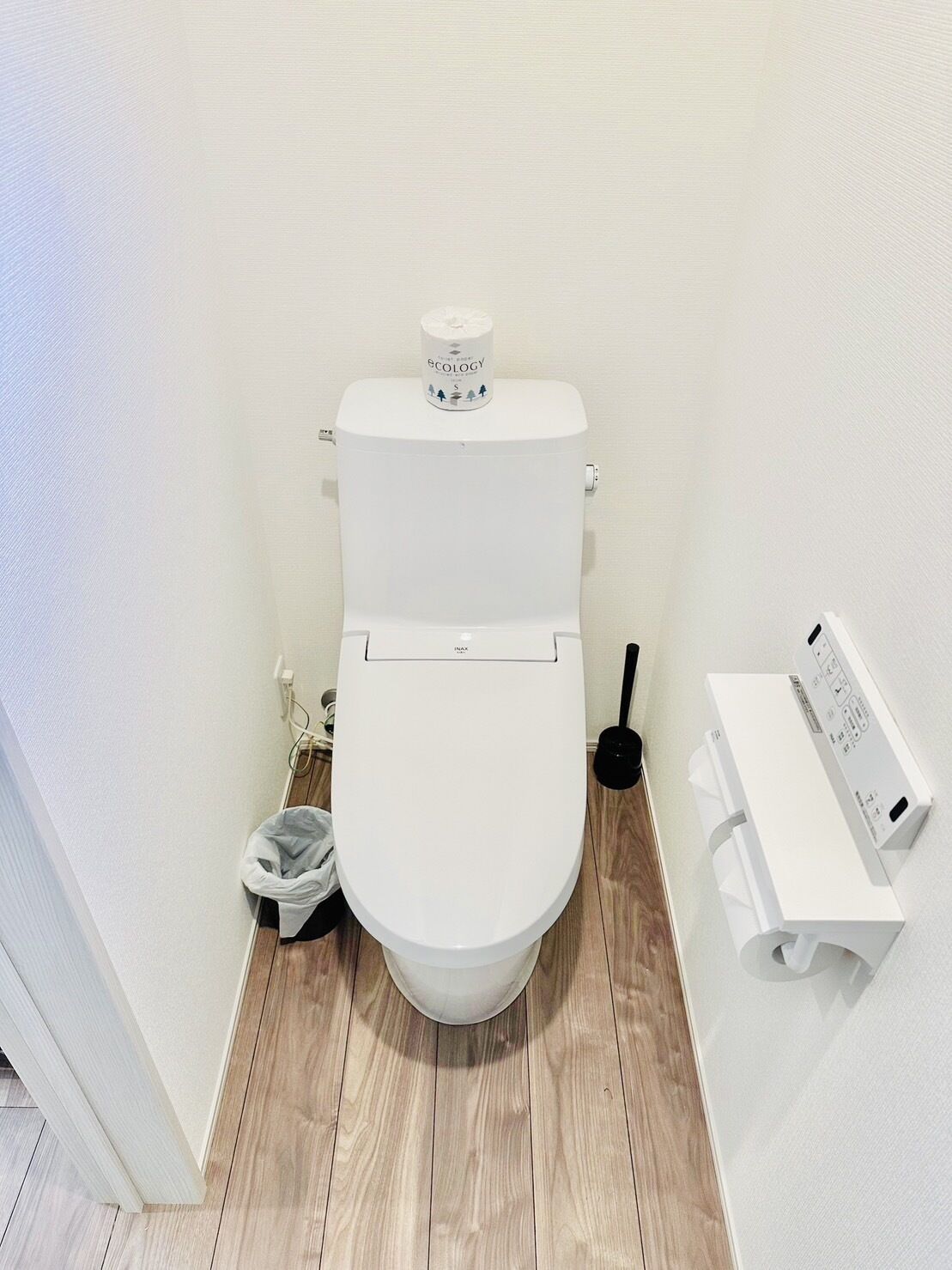 A white toilet with a toilet paper holder on the wall, a plunger, and a trash bin.