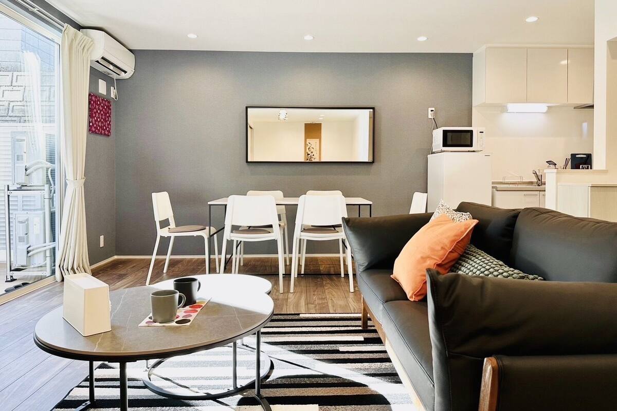 Una sala moderna con un sofá, una mesa de centro y un comedor con sillas. Hay una zona de cocina con un microondas y armarios.