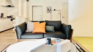 A modern living room with a black leather sofa, a coffee table with mugs and a tray, a kitchen area with a stove, and a door with an exit sign. - Megu Fuji Plus+ (Fujiyoshida)
